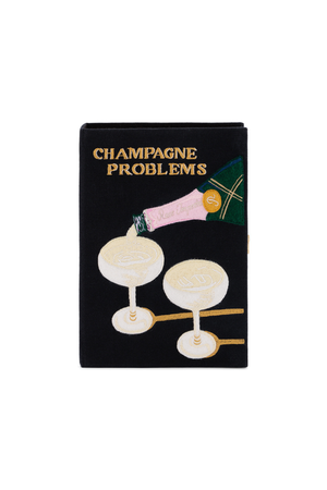 A black book clutch bag featuring a graphic illustration of a champagne bottle pouring into two champagne glasses, with the text 'CHAMPAGNE PROBLEMS' in gold lettering at the top.