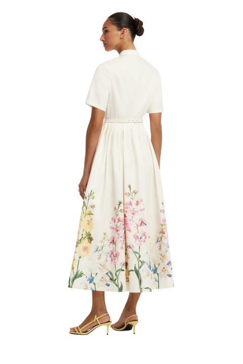 A woman shown from the back wearing a short-sleeve midi dress with a high mock neck, a thin waist belt, and a white skirt featuring a colorful floral print at the hem. She is wearing gold high-heeled sandals against a plain white background.