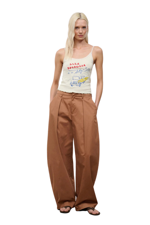A blonde model stands against a white background wearing a cream spaghetti-strap tank top with a colorful graphic print and high-waisted, wide-leg brown pants.