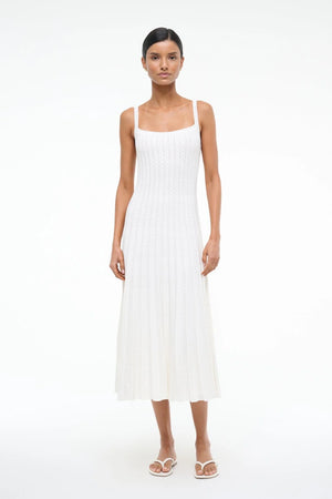 Woman wearing a white sleeveless midi dress that features eyelet trim detailing and a pleated skirt, paired with white sandals.