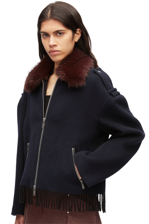 A model wearing a dark navy blue shearling-style jacket with a prominent dark brown faux-fur collar and black fringe along the hem, photographed against a clean white studio background.