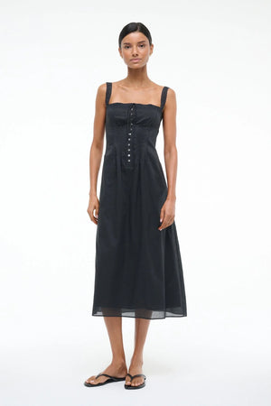 Woman wearing a black Victorian-inspired midi dress that features lace straps, corset-style detailing, and buttons down the front, paired with black sandals.