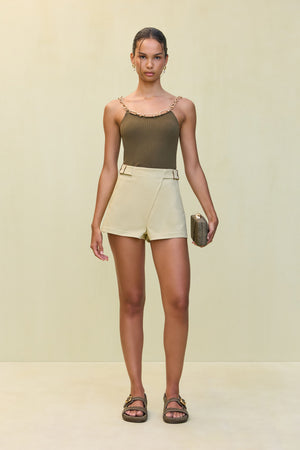 Woman wearing an olive green knit tank top that features a gold-toned chain detail for straps and across the neckline, paired with cream shorts and sandals.