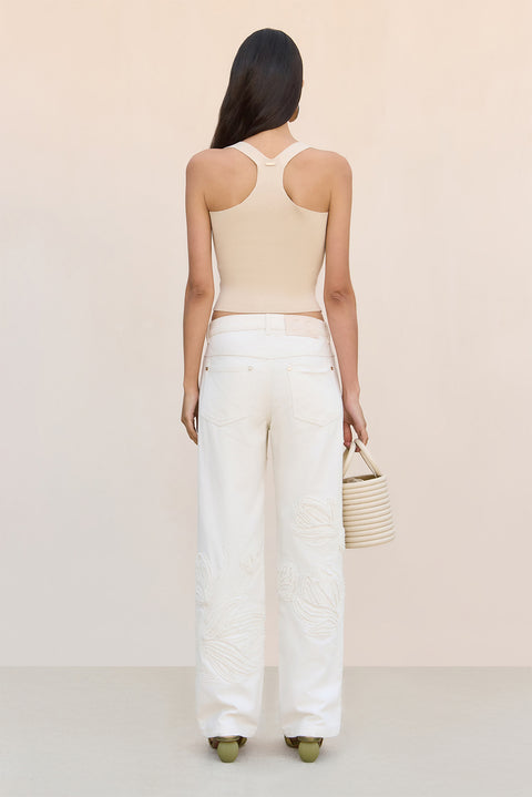 Back-facing shot of woman wearing a beige top and white pants holding a straw bag on a white background