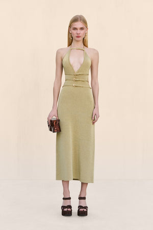 Woman wearing a gold knit midi dress that features a halter neck, deep V, and four buckle details across the bodice, paired with black accessories.