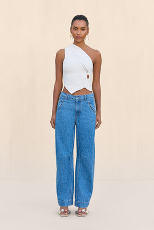 Woman wearing a white knit off the shoulder crop top that features a cutout at the rib cage and an asymmetrical hem, paired with blue wide leg jeans.