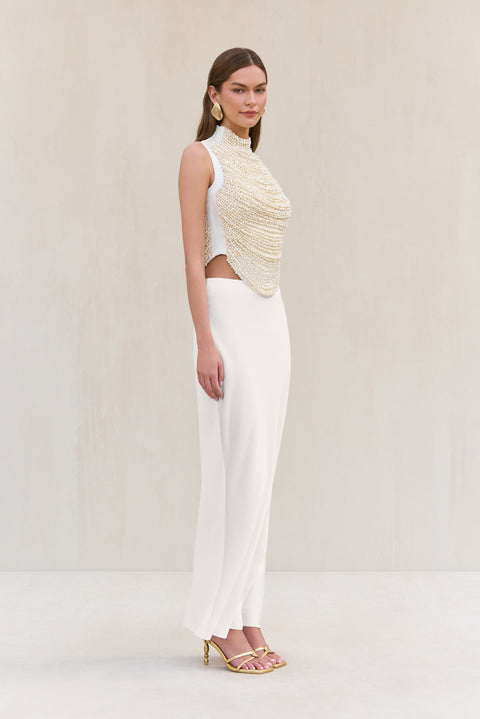 Side-facing woman wearing a cropped white tank top that features rows of pearl-beaded strands draped across the bodice, paired with a white maxi skirt.