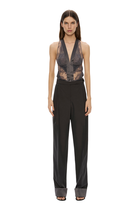 A model stands against a plain white background wearing a sleeveless black halter top with sheer lace panels and high-waisted wide-leg trousers.