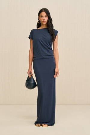 Woman wearing a slinky navy maxi dress that features an asymmetrical cap sleeve and draping throughout the bodice, paired with a navy handbag and gold sandals.