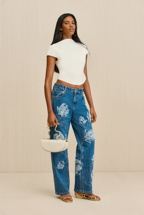 Woman wearing a pair of blue wash, wide leg jeans that features an embroidered floral design in lighter wash, paired with a white boatneck cropped t-shirt.