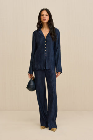 Woman wearing navy pleated pants, paired with a matching button down shirt, gold shoes, and a navy handbag.