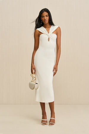 Woman wearing a white knit midi dress that features a twist and keyhole detail at the bust with both straps worn over the shoulders, paired with white sandals and a white handbag.
