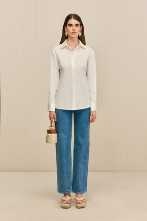 Woman wearing a white collared button down, paired with blue jeans, tan sandals, and a woven handbag.