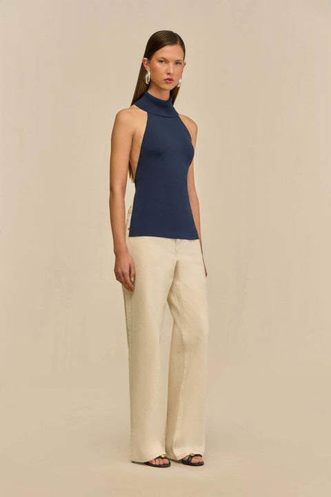 Woman wearing a navy sleeveless top and beige pants on a beige background from the side view.