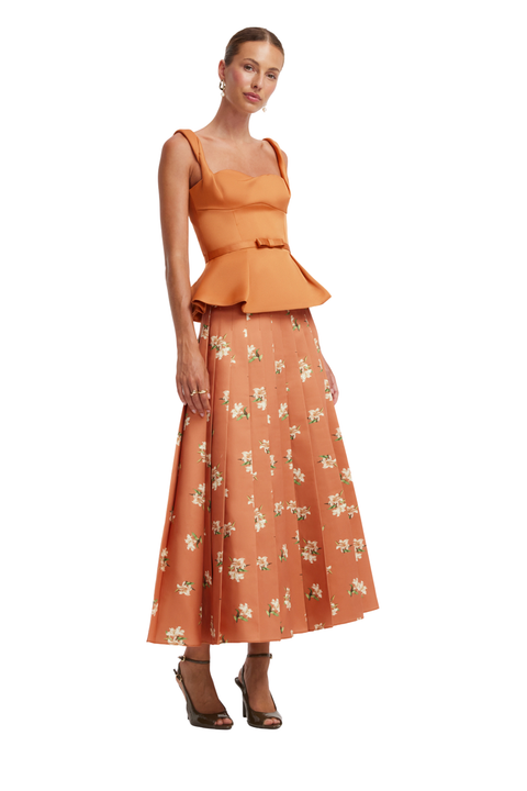 A woman modeling a two-piece outfit featuring a terracotta orange peplum top and a matching pleated midi skirt with a white floral print, set against a plain white background.