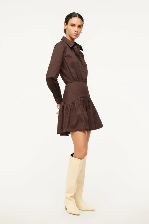 Side angle of a woman wearing a brown poplin mini dress that features long sleeves and a curved yoke detail at the hip, paired with tall, cream boots.
