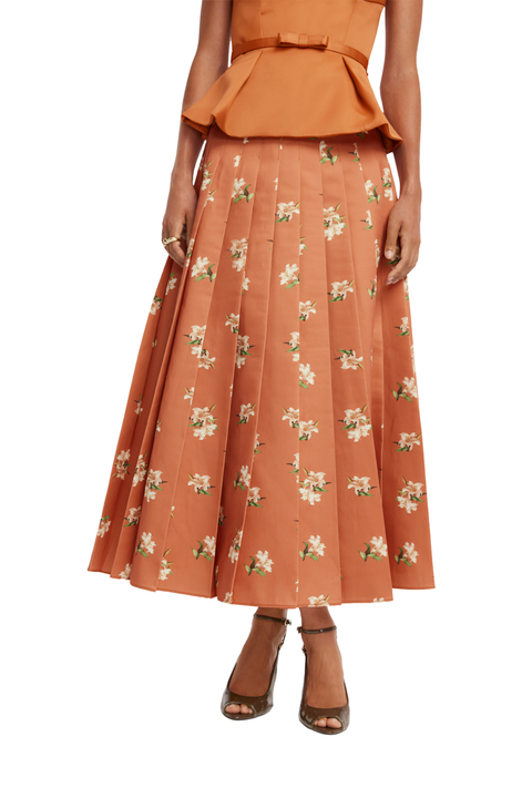A model wearing a rust-orange pleated midi skirt with a white floral print, paired with a matching peplum top and brown strappy heels, shown against a plain white background.