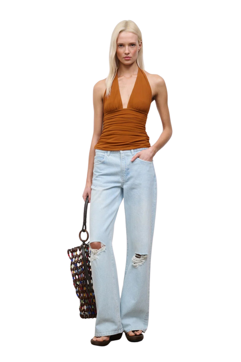 A blonde model stands against a white background wearing a brown halter top, light-wash distressed jeans, and sandals, while holding a woven brown and black handbag.