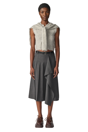 A model stands against a white studio background wearing a sleeveless textured light grey top with a polo collar, a dark gray pleated midi skirt with an asymmetrical hem and black belt, and brown lace-up shoes.