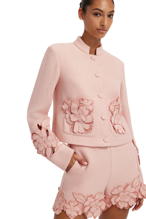A model wearing a light pink tweed jacket and matching shorts set, both decorated with large 3D floral appliques on the front and sleeves, set against a plain white background.