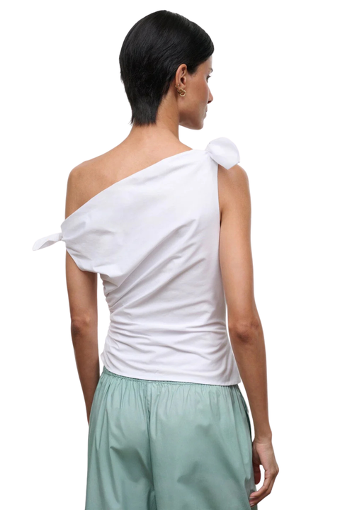 A model shown from the back wearing a white one-shoulder top with a knotted tie detail on the shoulder and light sage green pants, against a plain white background.