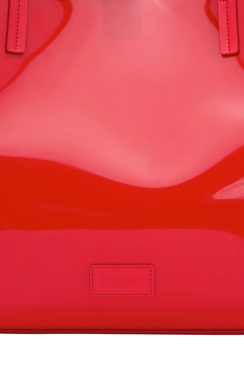 Close-up of a glossy bright pink-red patent leather bag with a small rectangular embossed logo patch near the bottom center.