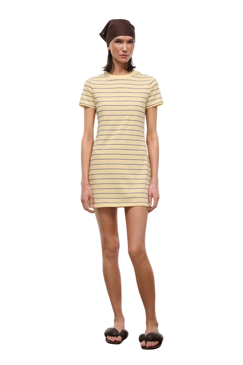 A model wearing a short-sleeve crewneck t-shirt dress with thin horizontal yellow stripes on a cream base, paired with a brown beanie and black slide sandals, against a plain white background.