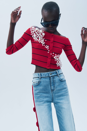 Person wearing a red cropped sweater with white pearl embellishments and light blue jeans on a white background