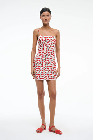 Woman wearing a white mini dress that features spaghetti straps, and square neck, and a red and green tomato pattern, paired with red sandals.