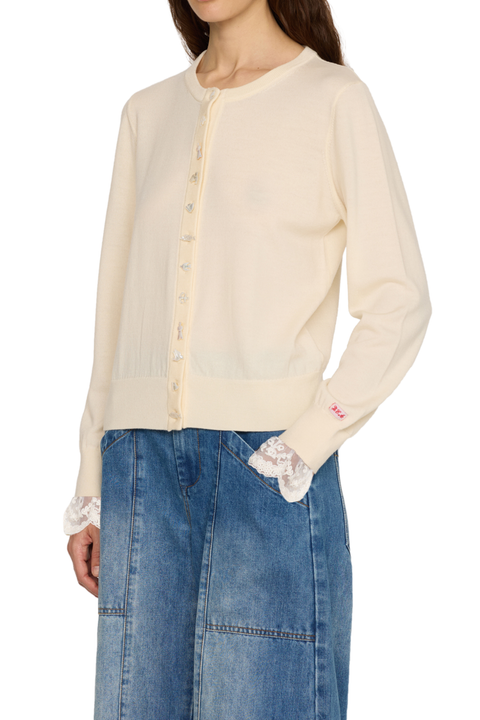 A model wearing a cream-colored long-sleeve knit cardigan with lace cuffs and a button-up front, paired with a blue denim skirt against a plain white background.