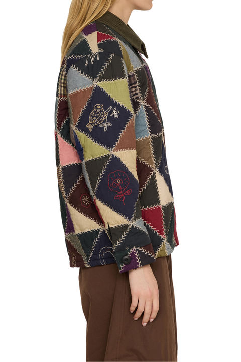 A woman shown from the side wearing a long-sleeve patchwork quilted jacket with geometric diamond panels in navy, beige, brown, green, pink, and red, paired with brown trousers, against a plain white background.