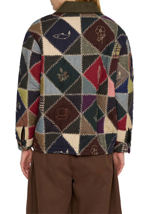 A person shown from the back wearing a quilted patchwork jacket with geometric diamond panels in navy, beige, red, green, and brown, paired with brown trousers, against a plain white background.