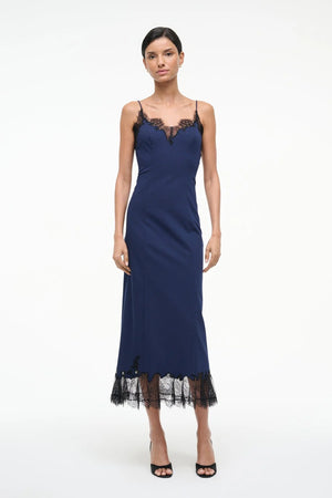 Woman wearing a navy midi slip dress that features black lace trim along the neckline and hem, paired with black kitten heels.