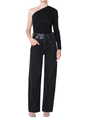 Person wearing a black, one-shoulder top and black wide-leg, high-rise leather and denim layered jeans on a white background right hand in pocket