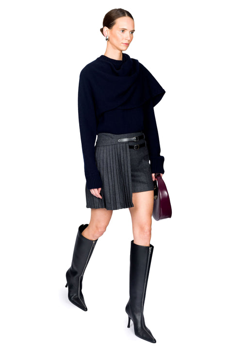Woman walking wearing a blue sweater, asymmetrical mini pinstripe skirt, and black knee-high boots on a white background