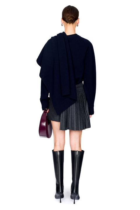 Woman facing backwards wearing a navy sweater, asymmetrical pleated skirt, and black boots on a white background