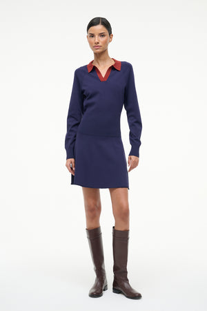Woman wearing a navy dress with a red collar and brown boots on a white background