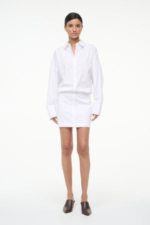 Woman wearing a long sleeve, white, mini shirt dress, paired with brown mules.
