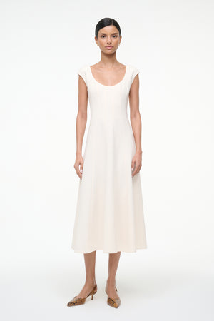 Woman wearing a cream, A-line midi dress that features a scoop neck, cap sleeves, and princess seams at the waist, paired with kitten heels.