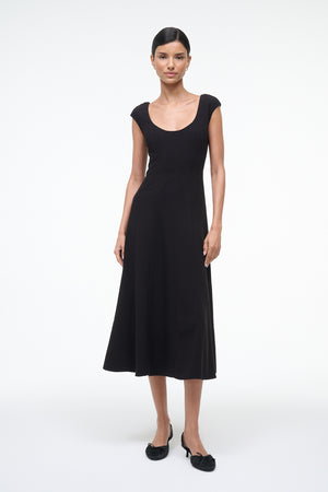Woman wearing a black, A-line, midi dress that features a scoop neck, princess seams at the waist, and cap sleeves, paired with black kitten heels.