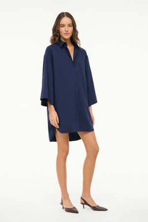 Woman wearing a navy shirt dress that features a 3/4 length sleeve, collar, and button down detail, paired with brown slingbacks.