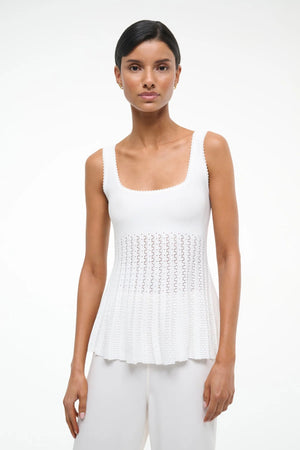 Close up of a woman wearing a white knit tank top that features a square neck and sheer, pleated bodice, paired with white pants.
