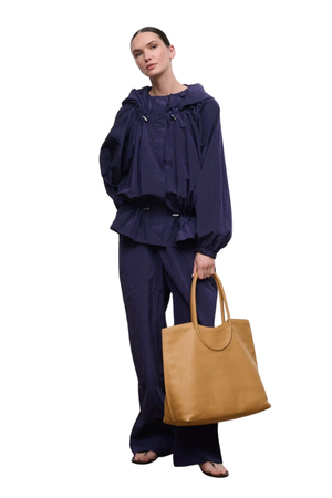 A model wearing a dark navy blue hooded jacket and matching wide-leg pants, holding a large tan leather tote bag against a solid white background.