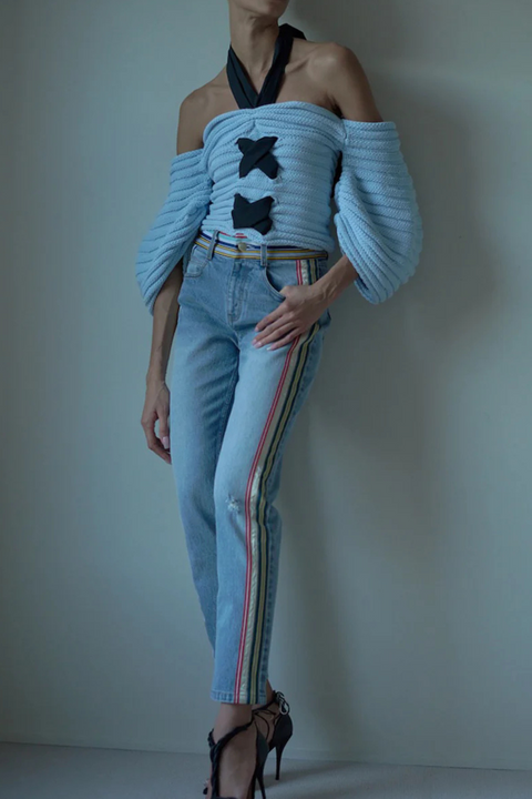 Woman wears skinny leg denim in light wash with striped grosgrain detail at the waist and down the side of the leg with blue knitted top and heels.