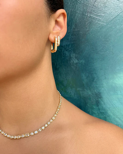 A woman wearing a rectangular gold hoop earring that features inset baguette diamonds, stacked with a smaller gold hoop that features scattered diamonds, paired with a multi shape tennis necklace.