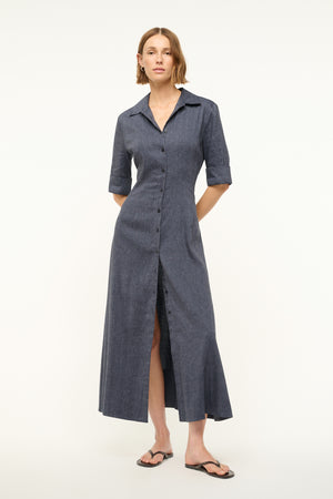 Woman wearing a navy linen maxi dress that features a cuffed half sleeve, collar, and button down detail, paired with black sandals.