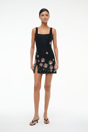 Woman wearing a sleeveless beaded mini dress that features a square neck and side slit, designed with pink dahlias on a black background, paired with black heels.