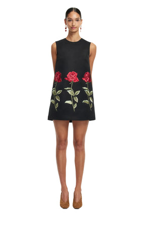 Woman wears a black sleeveless mini dress with red floral embroidery on a white background