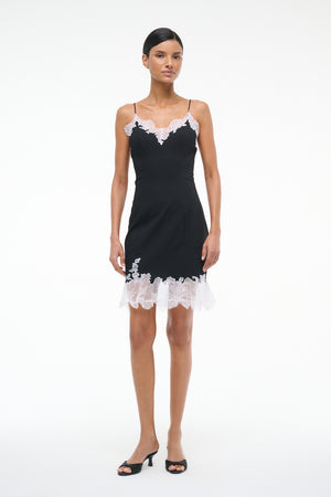 Woman wearing a black mini slip dress with white lace trim on the neckline and hem, paired with black kitten heels.