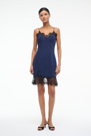 Woman wearing a navy blue mini slip dress with black lace trim on the neckline and hem, paired with black kitten heels.